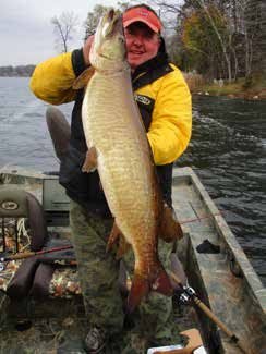 A smiling angler wearing a yellow and black jacket and red cap holds up a massive muskellunge (muskie) in a small fishing boat on a lake, surrounded by fall trees.