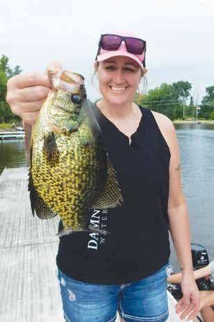 A smiling woman wearing a sleeveless black shirt, denim shorts, and pink sunglasses holds up a large crappie on a dock beside a calm lake with trees and boats in the background.