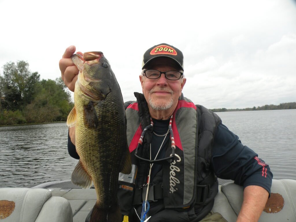An older man wearing glasses, a cap, and a life vest sits in a boat holding a largemouth bass upright by the mouth. A calm lake and tree-lined shoreline stretch out behind him under an overcast sky.
