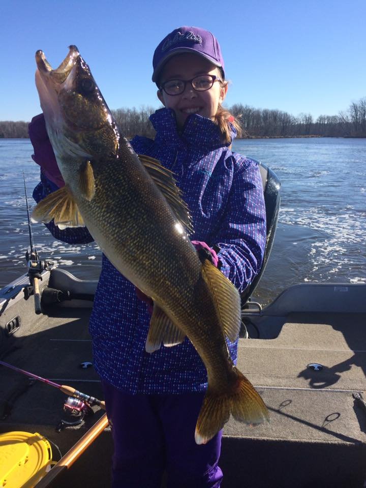 A young angler dressed in a purple jacket and hat proudly holds up a large walleye while standing in a boat on a cold, sunny day with open water and bare trees in the background.