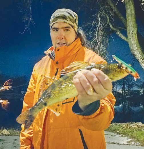 A person wearing an orange jacket and a camo headwrap holds a walleye toward the camera at night. Houses and trees are visible in the dimly lit background near a calm waterfront.