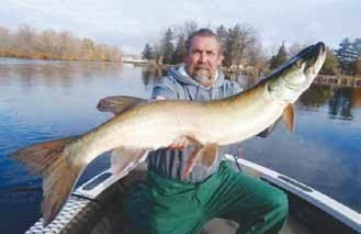 A man sitting at the front of a small boat holds a large muskellunge horizontally toward the camera. Calm water and autumn trees line the background under a bright sky.
