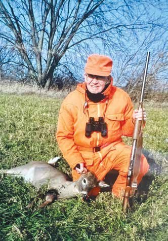 A person dressed in blaze-orange hunting gear kneels in a grassy field beside a harvested deer, holding a rifle upright and resting a hand on the deer’s head. Leafless trees and a bright blue sky are visible in the background.
