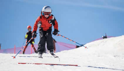 A young skier in an orange jacket and helmet glides downhill with ski poles extended, approaching a small jump on a sunny, snow-covered slope. Another skier stands in the background.