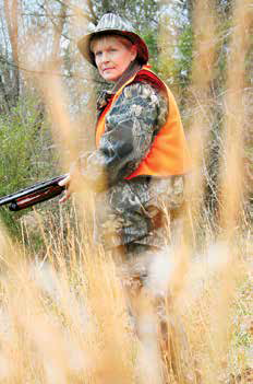 Hunter wearing an orange safety vest holding a shotgun in tall grass.