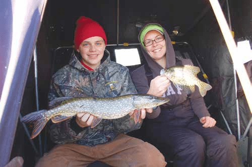 Two ice anglers sitting inside an ice shelter holding a northern pike and a crappie.