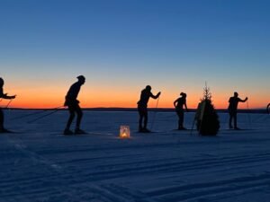 Silhouettes of several cross-country skiers gliding across a snowy landscape at sunset, with a glowing lantern and a small evergreen tree in the foreground against a vivid orange and blue sky.