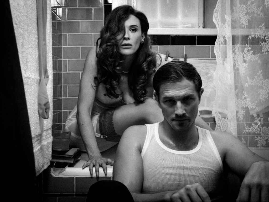 Black-and-white promotional image of a tense couple in a small bathroom, with a woman leaning over the edge of a bathtub behind a brooding man seated in front.
