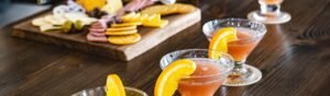 Three pink-orange cocktails garnished with fresh orange slices sit on a wooden bar top, with a blurred charcuterie board of cheeses, crackers, olives, and sliced meats in the background.